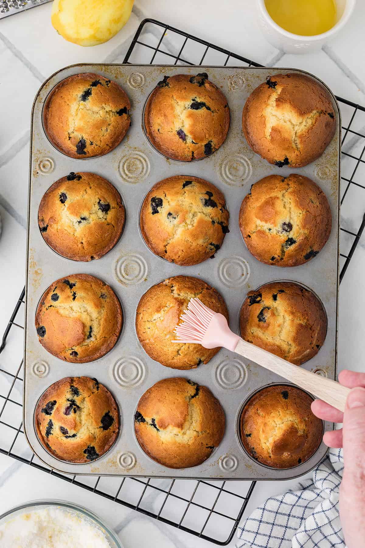 A hand brushes the tops of freshly baked blueberry muffins in a muffin tin with melted butter on a cooling rack.