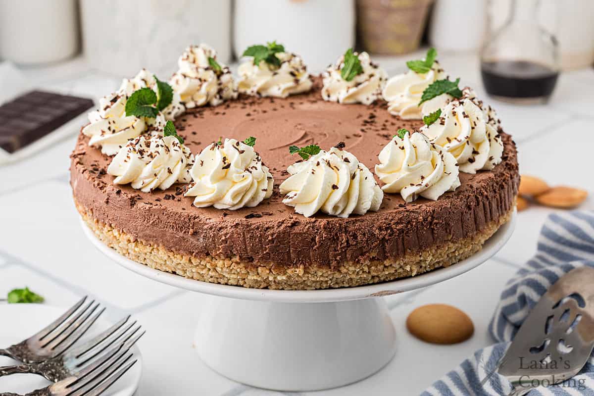 A chocolate cheesecake with a cookie crumb crust, topped with whipped cream, chocolate shavings, and mint leaves, displayed on a white cake stand.