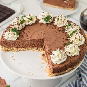 A chocolate cheesecake with a slice missing, topped with whipped cream, chocolate shavings, and mint leaves, displayed on a white cake stand.