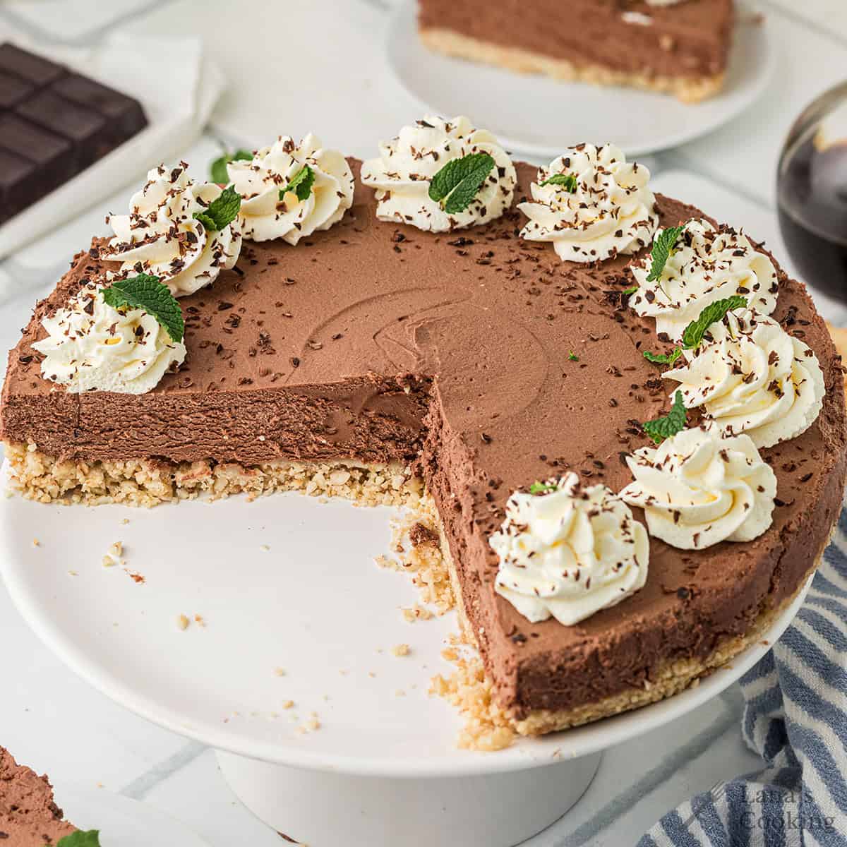 A chocolate cheesecake with a slice missing, topped with whipped cream, chocolate shavings, and mint leaves, displayed on a white cake stand.