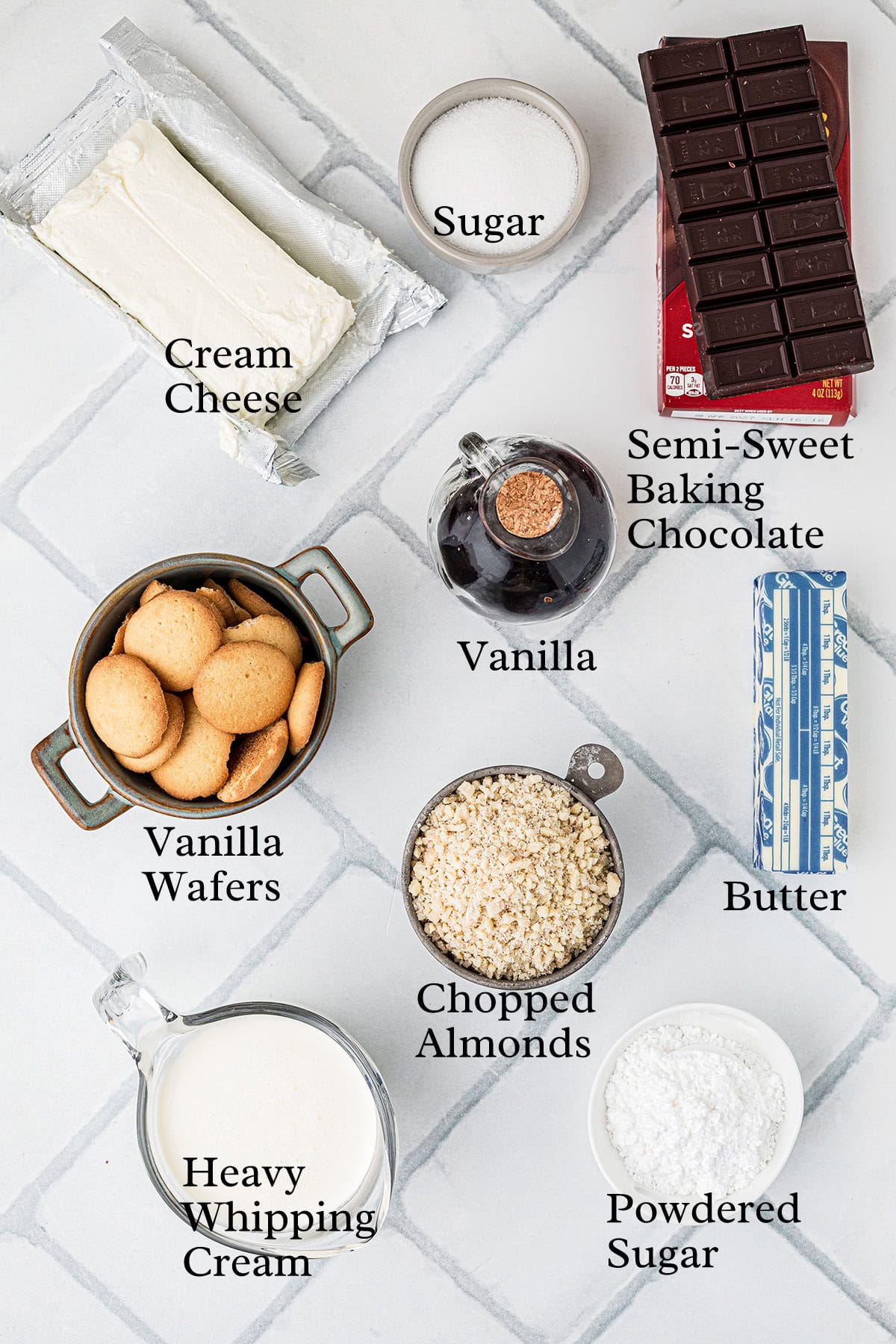 Ingredients for the recipe arranged on a white tiled surface, including cream cheese, sugar, chocolate, vanilla wafers, vanilla, almonds, butter, heavy cream, and powdered sugar.