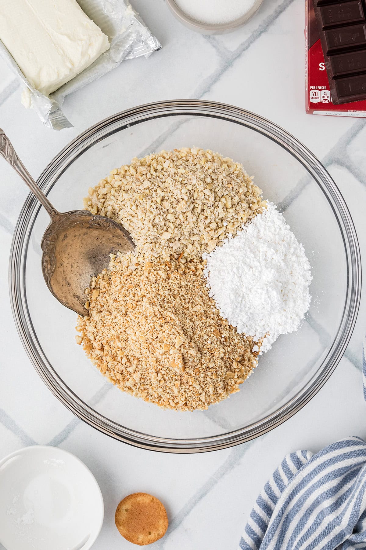 A glass bowl contains chopped almonds, crushed vanila wafers, powdered sugar, with a metal spoon resting inside. Cream cheese, chocolate, and other ingredients are nearby on a marble surface.
