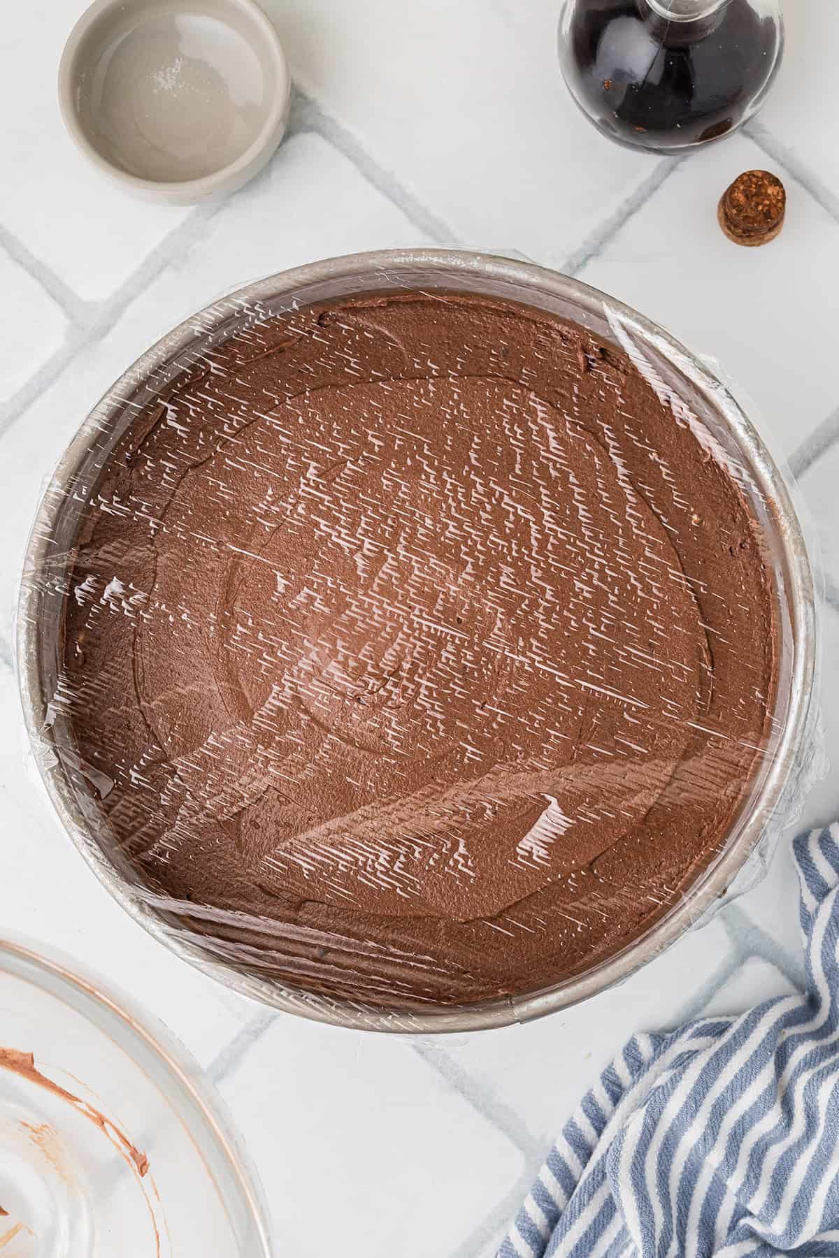 A round pan filled with chocolate mousse covered with plastic wrap, placed on a white tiled surface next to a striped towel and a small bowl.