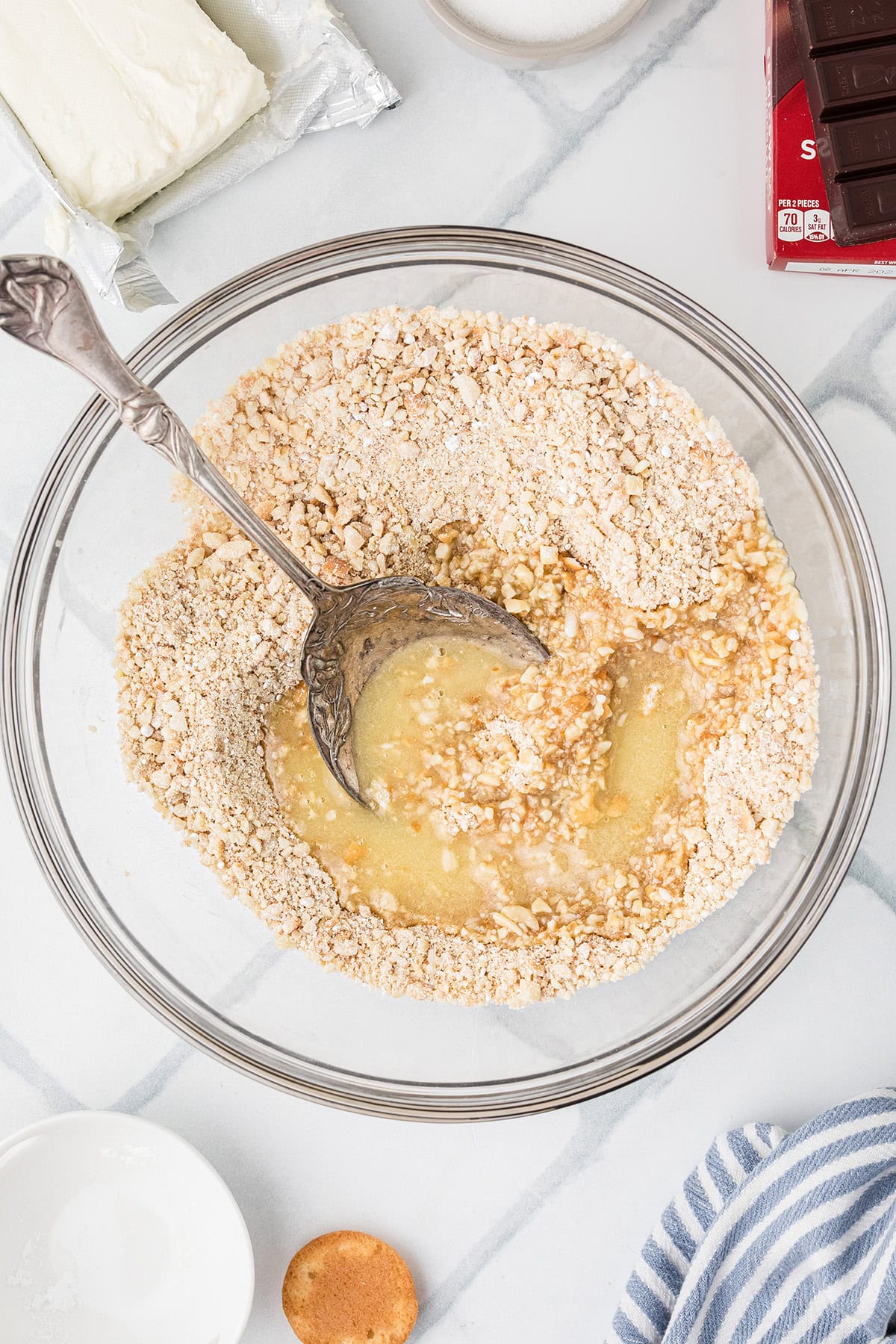 A bowl containing crushed cookies and almonds and melted butter being mixed with a large spoon, surrounded by cream cheese, chocolate, and kitchen utensils on a marble surface.