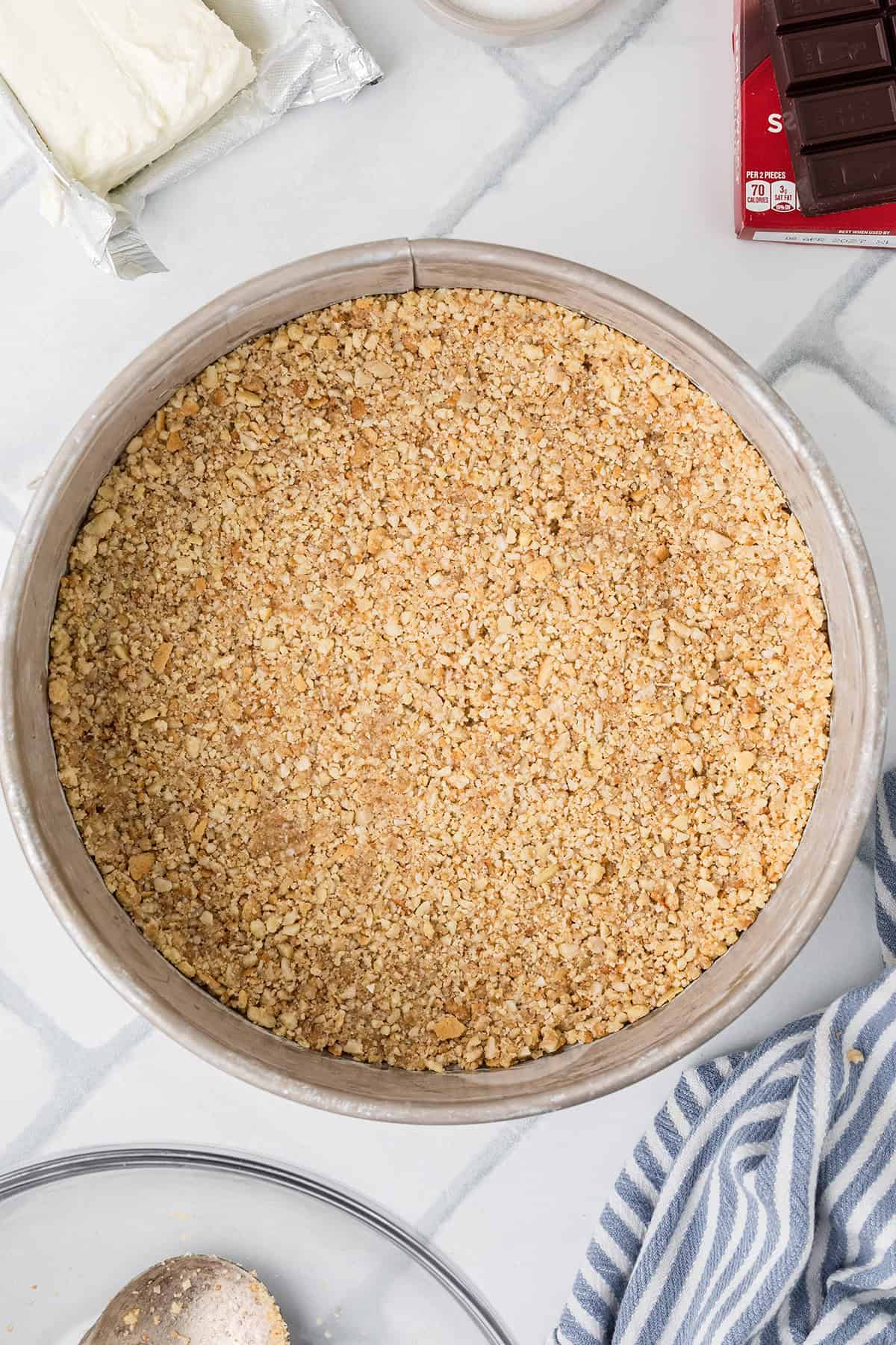 A springform pan filled with an even layer of almond and cookie crust, surrounded by baking ingredients on a marble countertop.