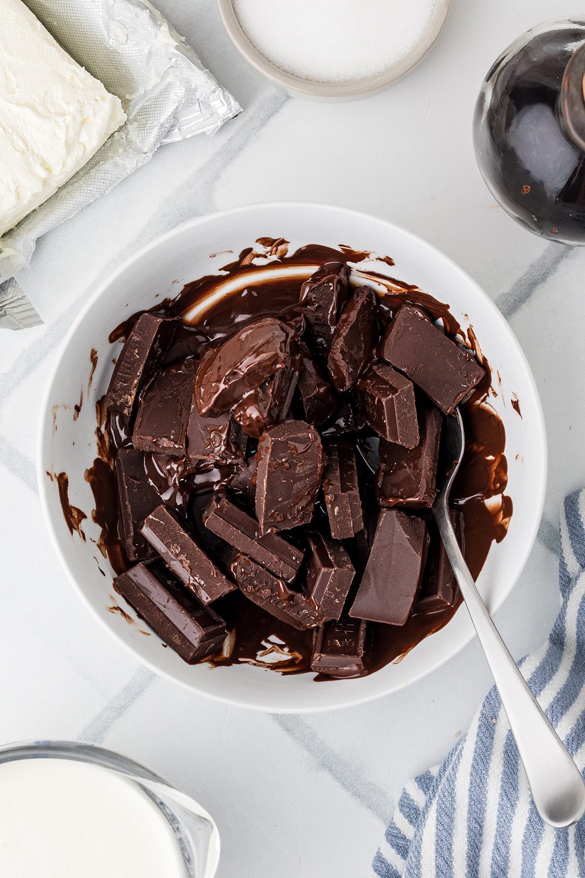 A white bowl filled with partially melted chocolate chunks and a spoon, surrounded by cream, cream cheese, sugar, and a striped cloth.