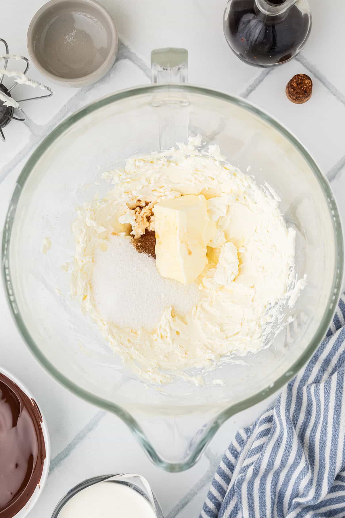 A glass mixing bowl containing cream cheese, butter, sugar, and vanilla extract sits on a counter with baking ingredients nearby.