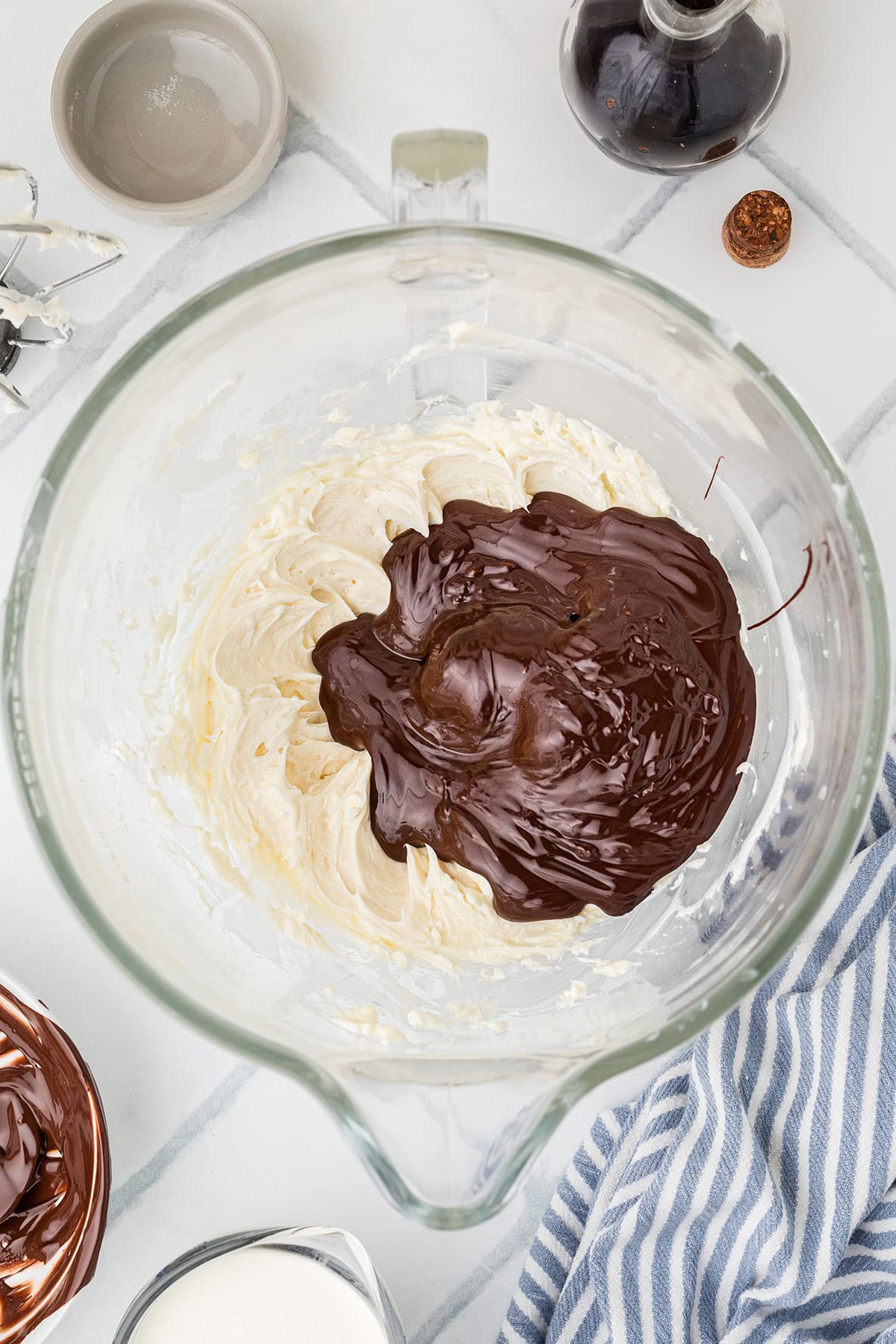 A glass mixing bowl with creamy batter and melted chocolate, surrounded by baking tools and ingredients on a marble countertop.
