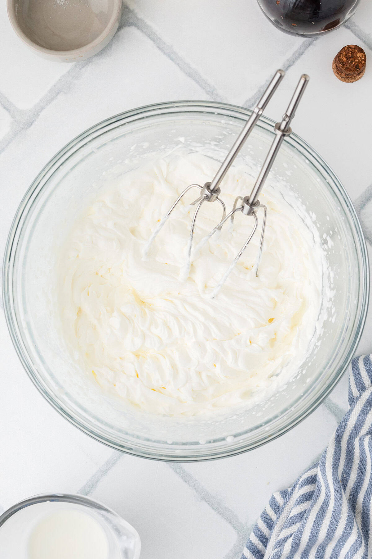 A glass bowl filled with whipped cream and an electric hand mixer resting inside, on a white countertop with a striped towel and small dishes nearby.