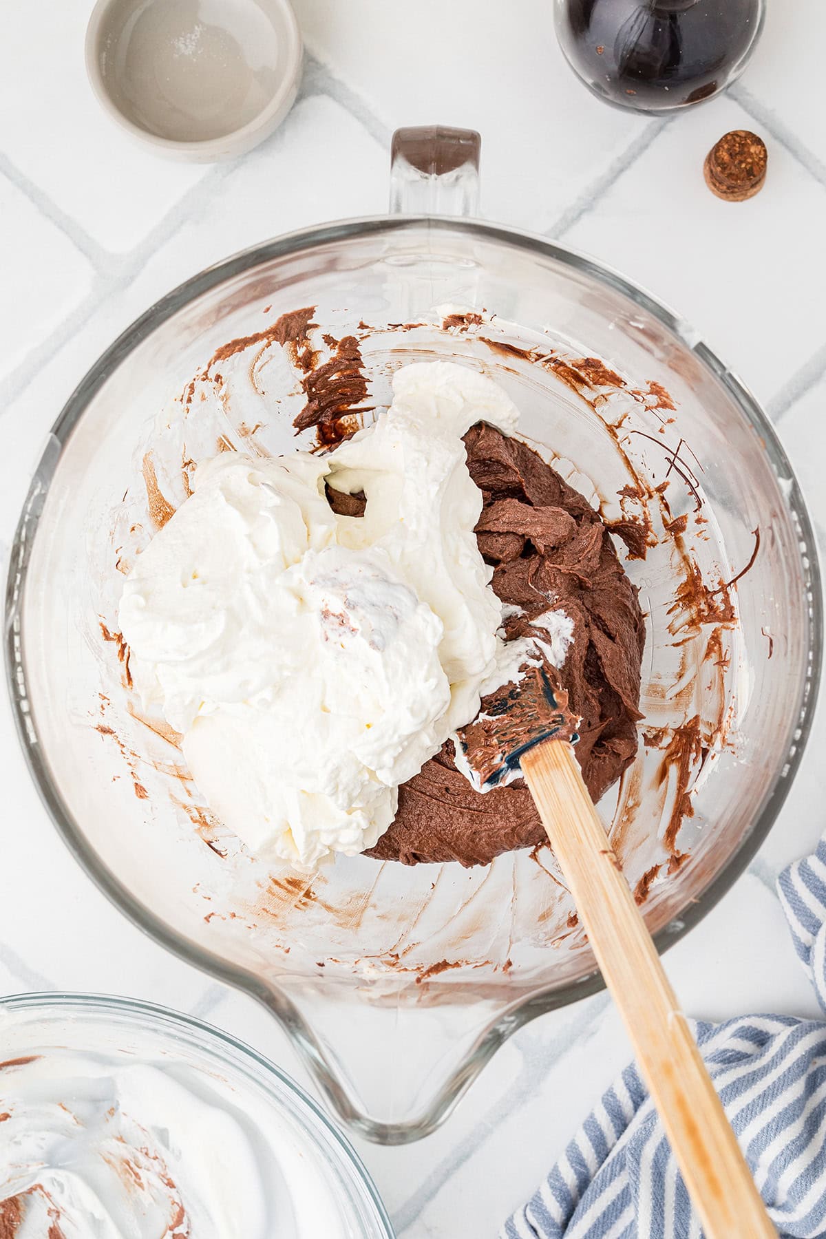 A glass mixing bowl contains whipped cream and chocolate mixture being combined with a spatula on a white tiled surface; other ingredients sit nearby.