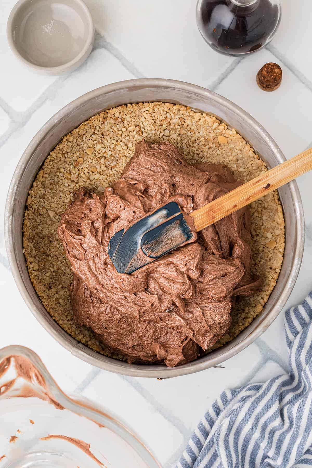 A baking pan with a nutty crust base is being filled with chocolate mixture, spread by a spatula. Nearby are a striped towel, a bowl, and a bottle of vanilla extract.