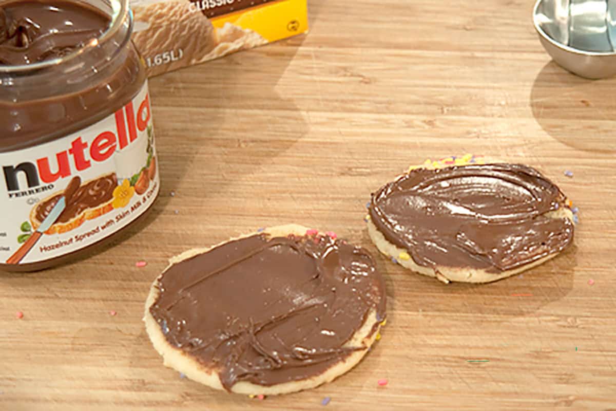 Two round cookies on a wooden surface, each topped with a layer of Nutella spread; an open Nutella jar and measuring spoon are nearby.