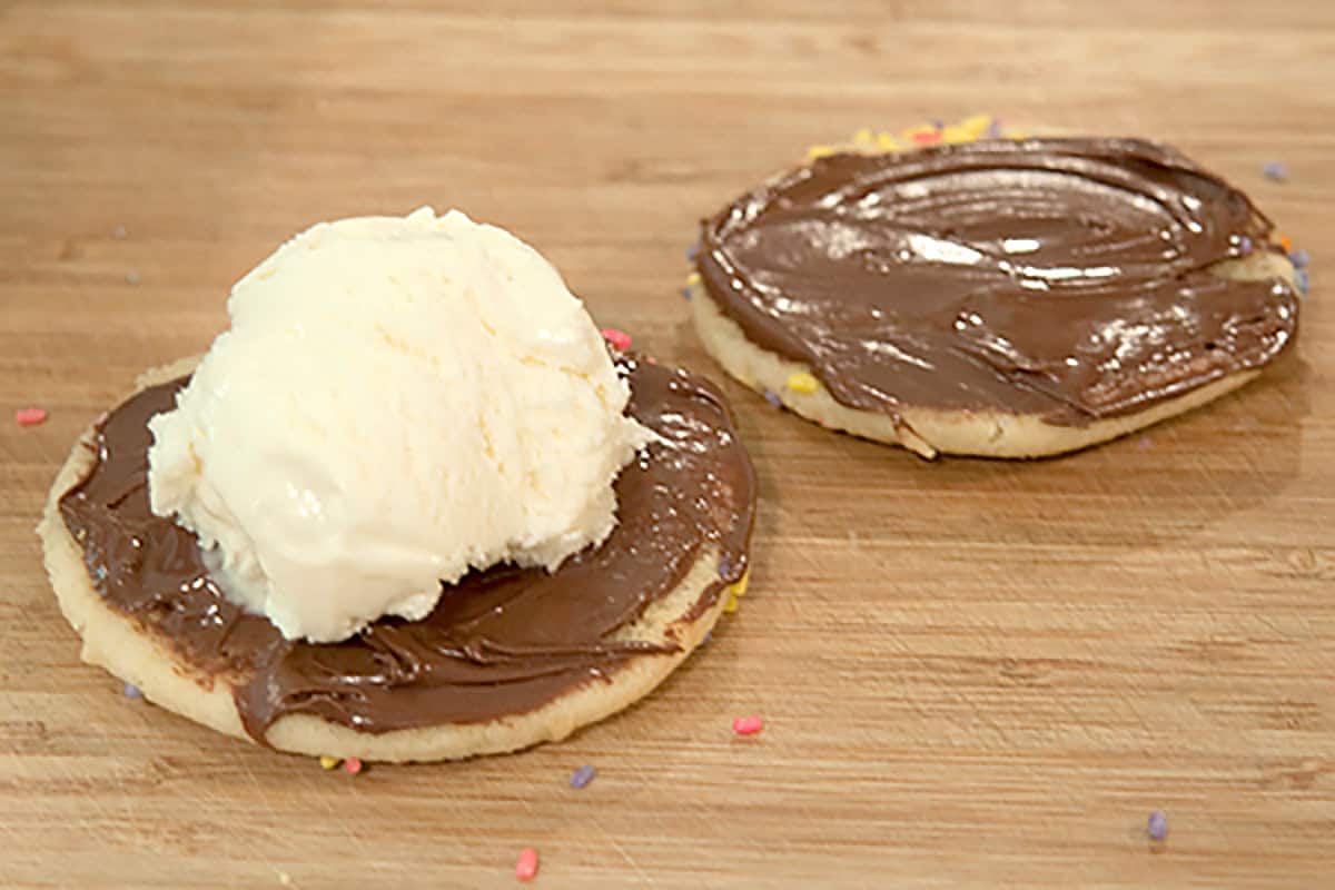 Two sugar cookies with Nutella spread; one has a scoop of vanilla ice cream on top. Both are on a wooden surface with a few scattered sprinkles.