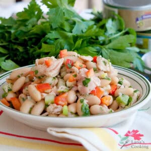 A bowl of white bean salad with tuna, chopped red bell pepper, celery, and parsley sits on a striped cloth, with fresh parsley and a can in the background.