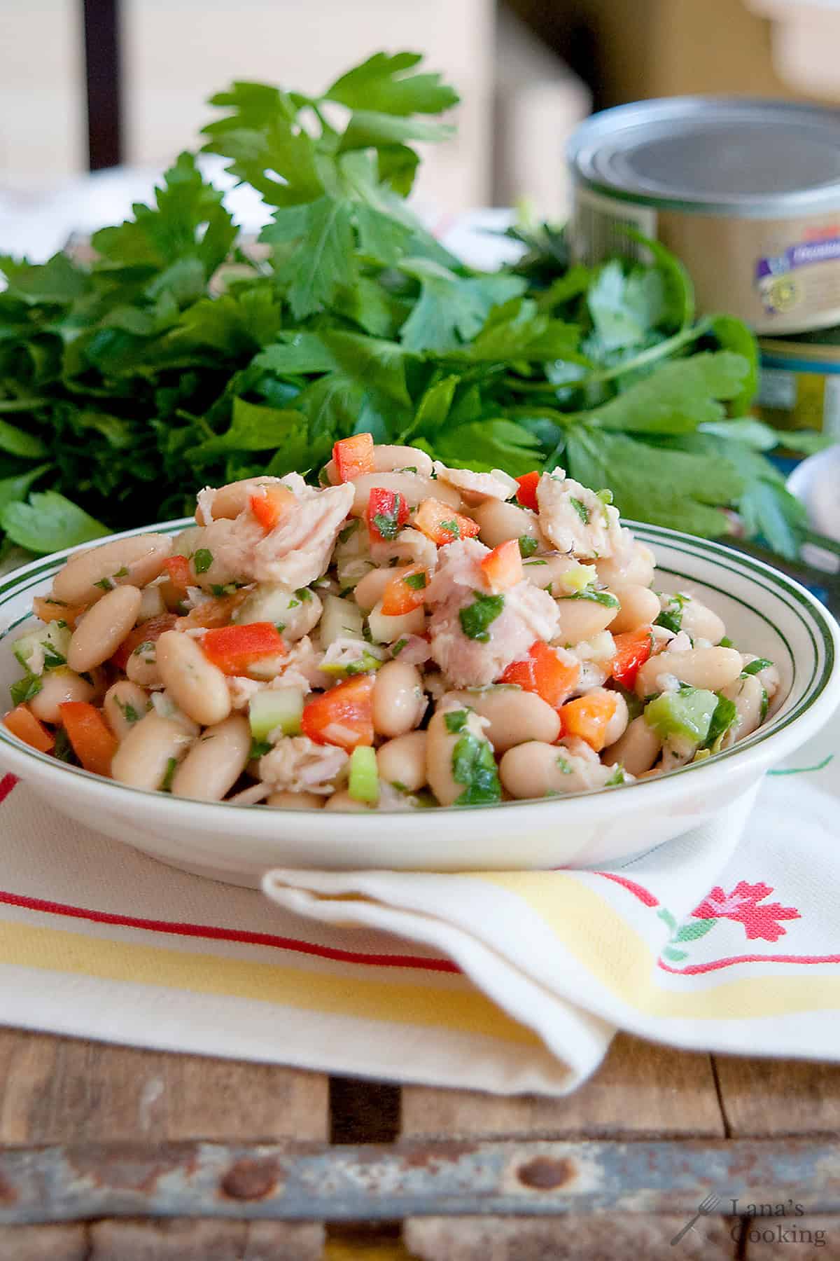 A bowl of white bean and tuna salad with chopped vegetables sits on a napkin, with fresh parsley and a can in the background.