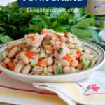 A bowl of cannellini bean and tuna salad with chopped vegetables, placed on a cloth with fresh herbs behind.
