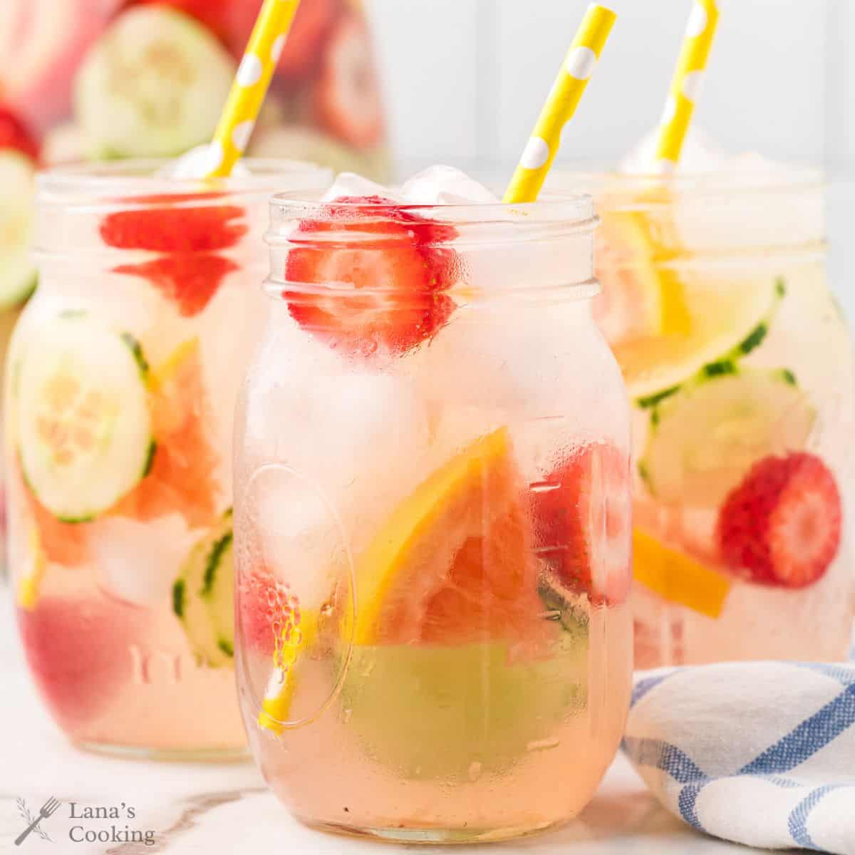 Three mason jars of infused water with ice, strawberries, pink grapefruit, cucumber, and yellow striped straws.