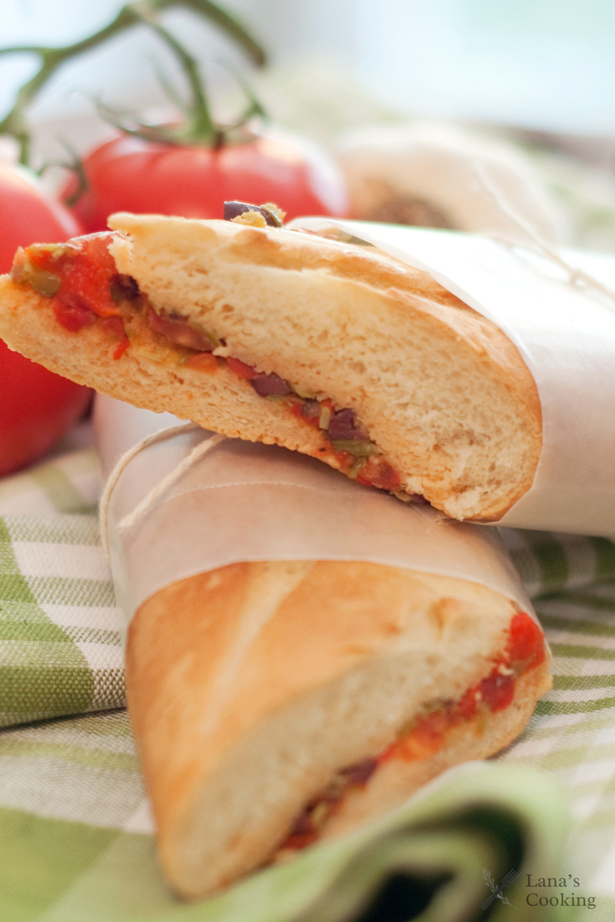 A sliced baguette sandwich with tomato, vegetables, and sauce, wrapped in parchment paper, is displayed on a green and white checkered cloth with fresh tomatoes in the background.