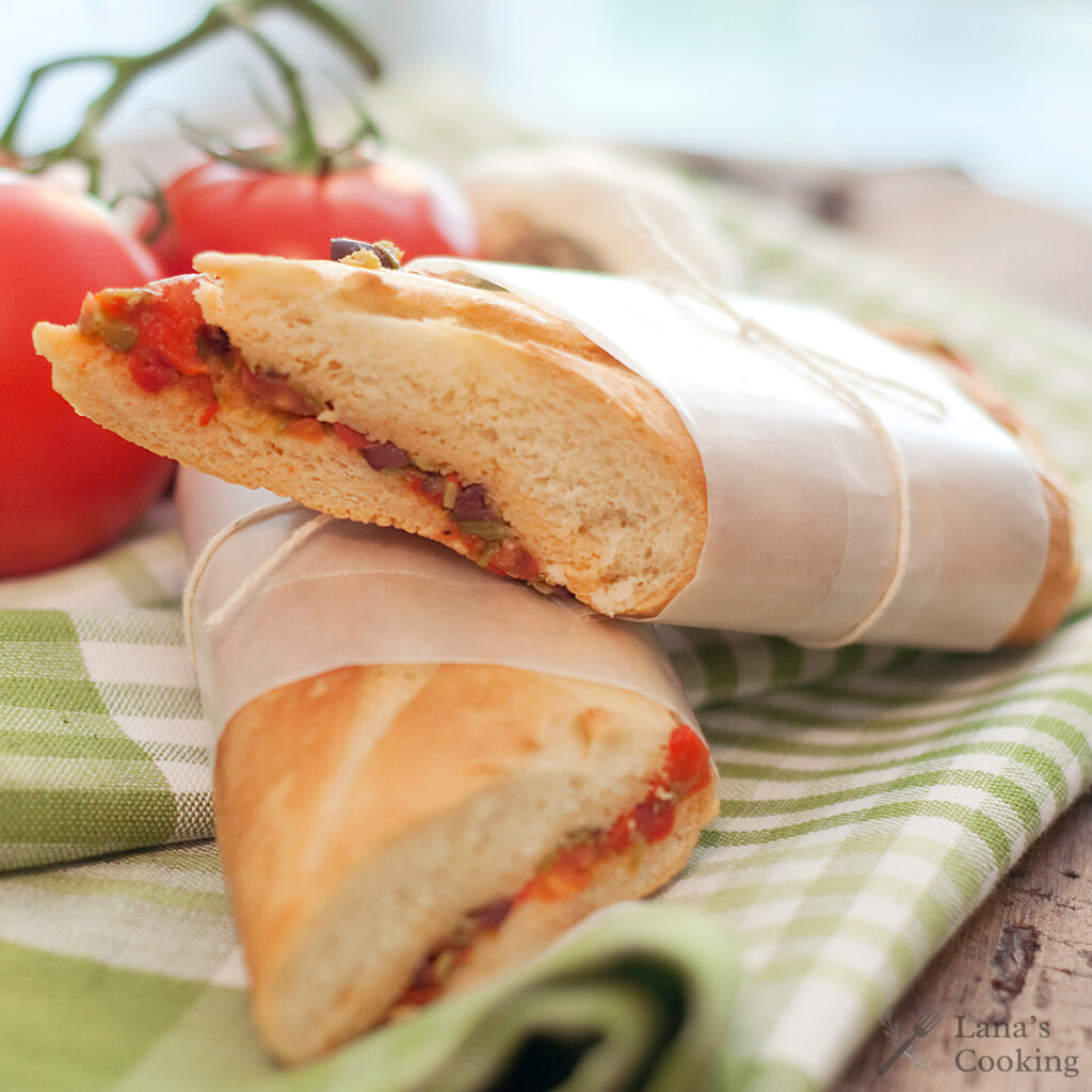 A baguette sandwich filled with tomato, onion, and green peppers sits on a green checked cloth, wrapped in parchment paper. Fresh tomatoes are in the background.