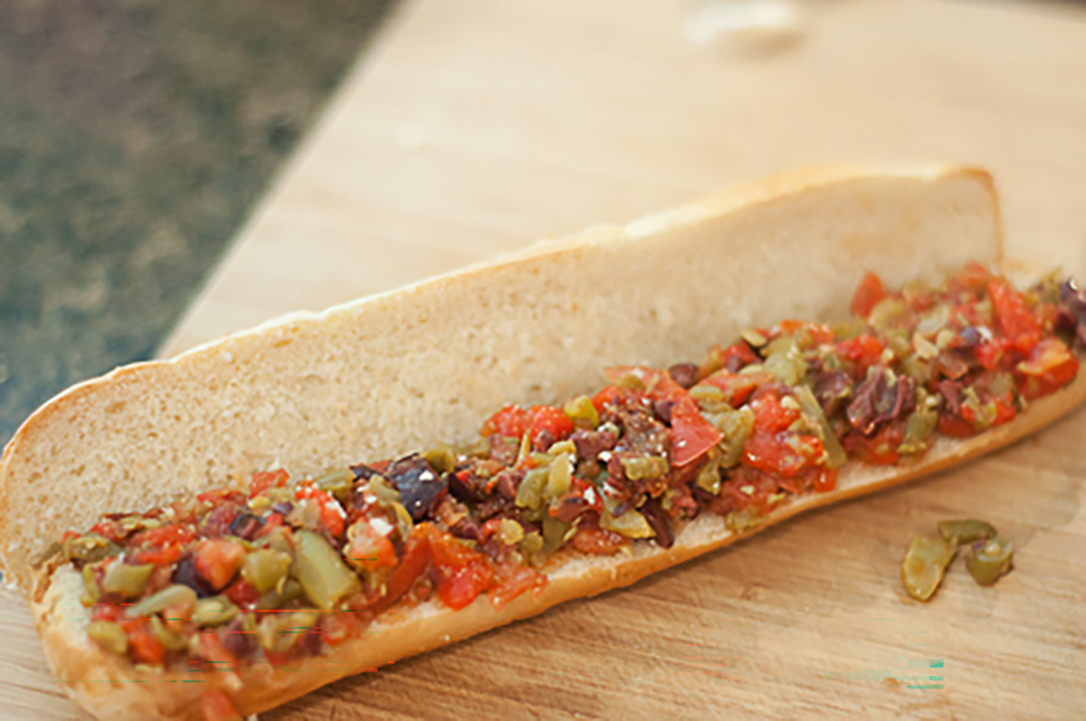 A sandwich roll on a wooden board filled with a chopped mixture of green and black olives, tomatoes, and other vegetables.