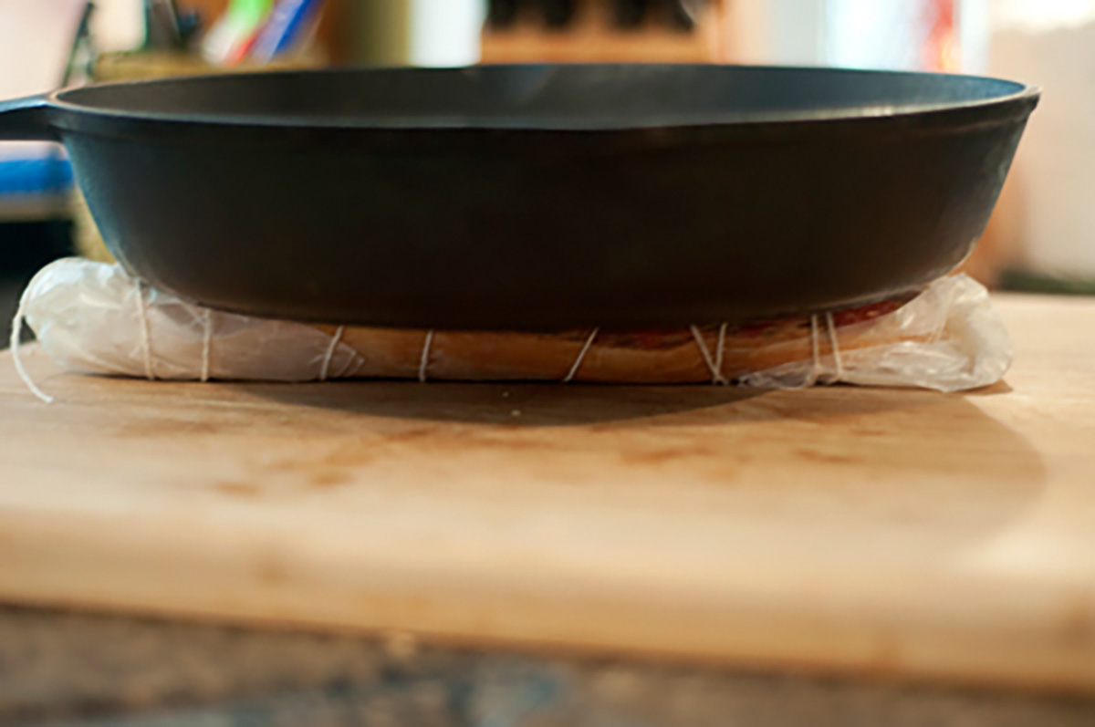 A cast iron skillet presses down on a wrapped sandwich on a wooden cutting board.