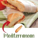 Two halves of a Mediterranean pressed sandwich are stacked on a green checkered cloth, with tomatoes and herbs visible in the background.