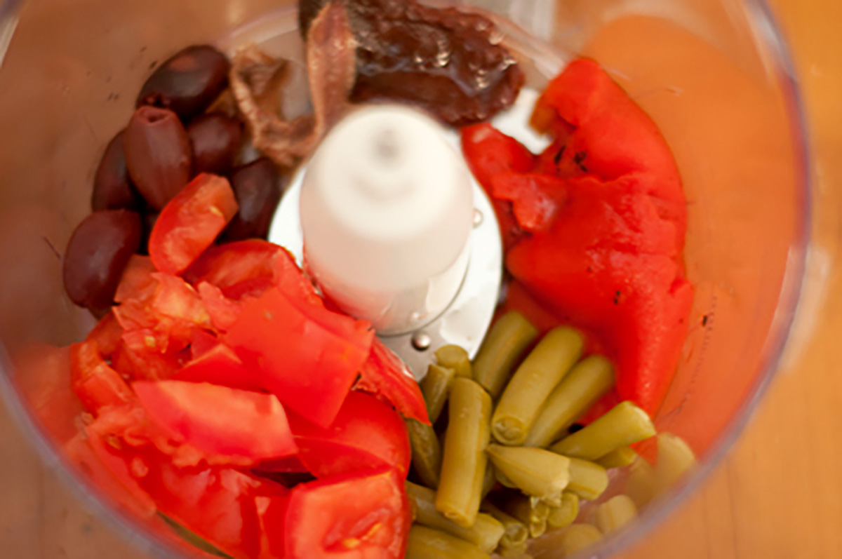 Chopped tomatoes, green beans, red peppers, olives, and anchovies inside a food processor bowl.