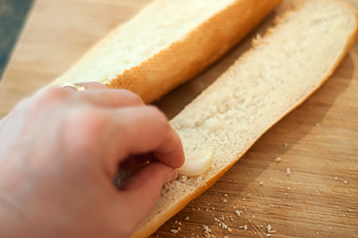 A hand rubbing a garlic clove on the inside of a sliced baguette placed on a wooden cutting board.