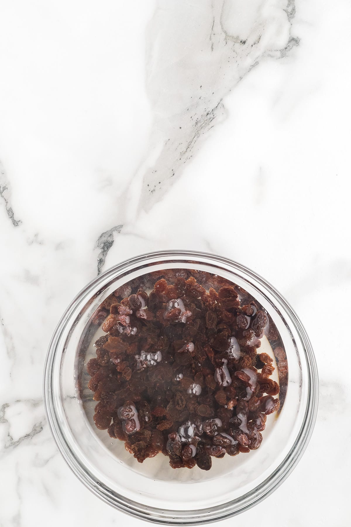 A glass bowl of raisins soaking in water on a white marble surface.