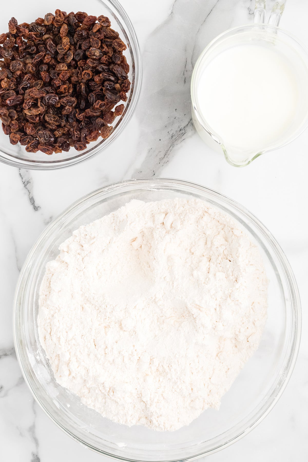 Bowl of flour, bowl of raisins, and a measuring cup of milk on a marble countertop.