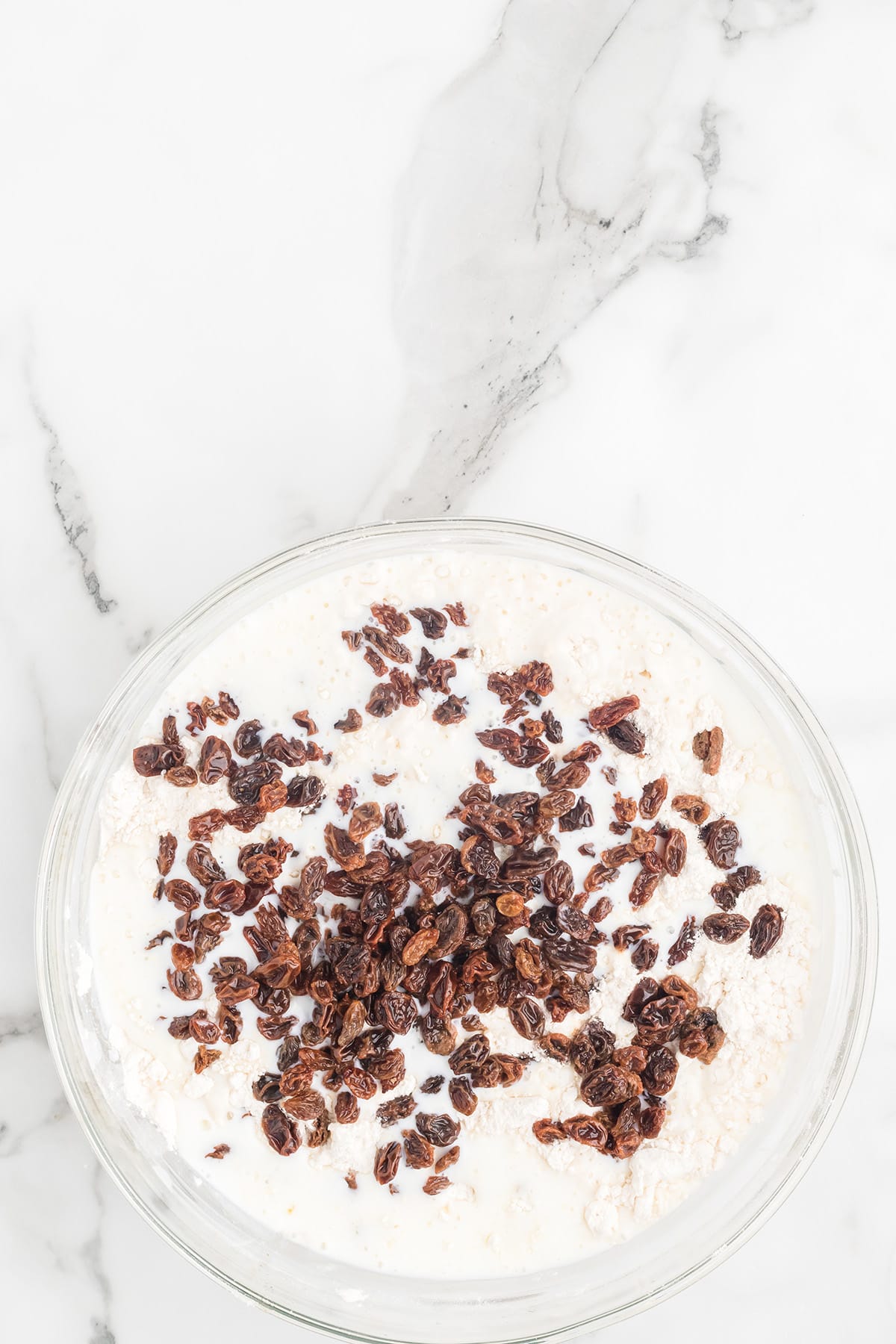 A glass bowl with milk, flour, and raisins on a white marble surface.