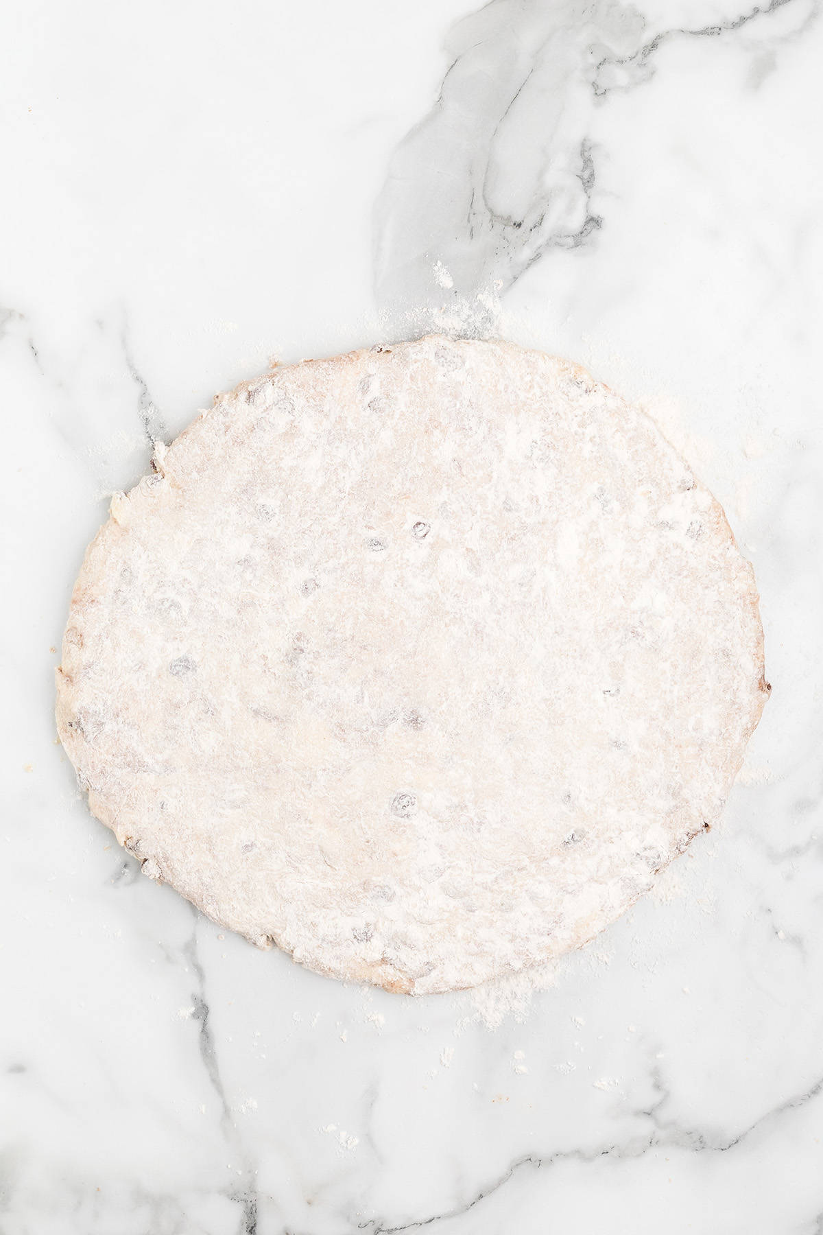 Round, floured dough is rolled out on a white marble surface.