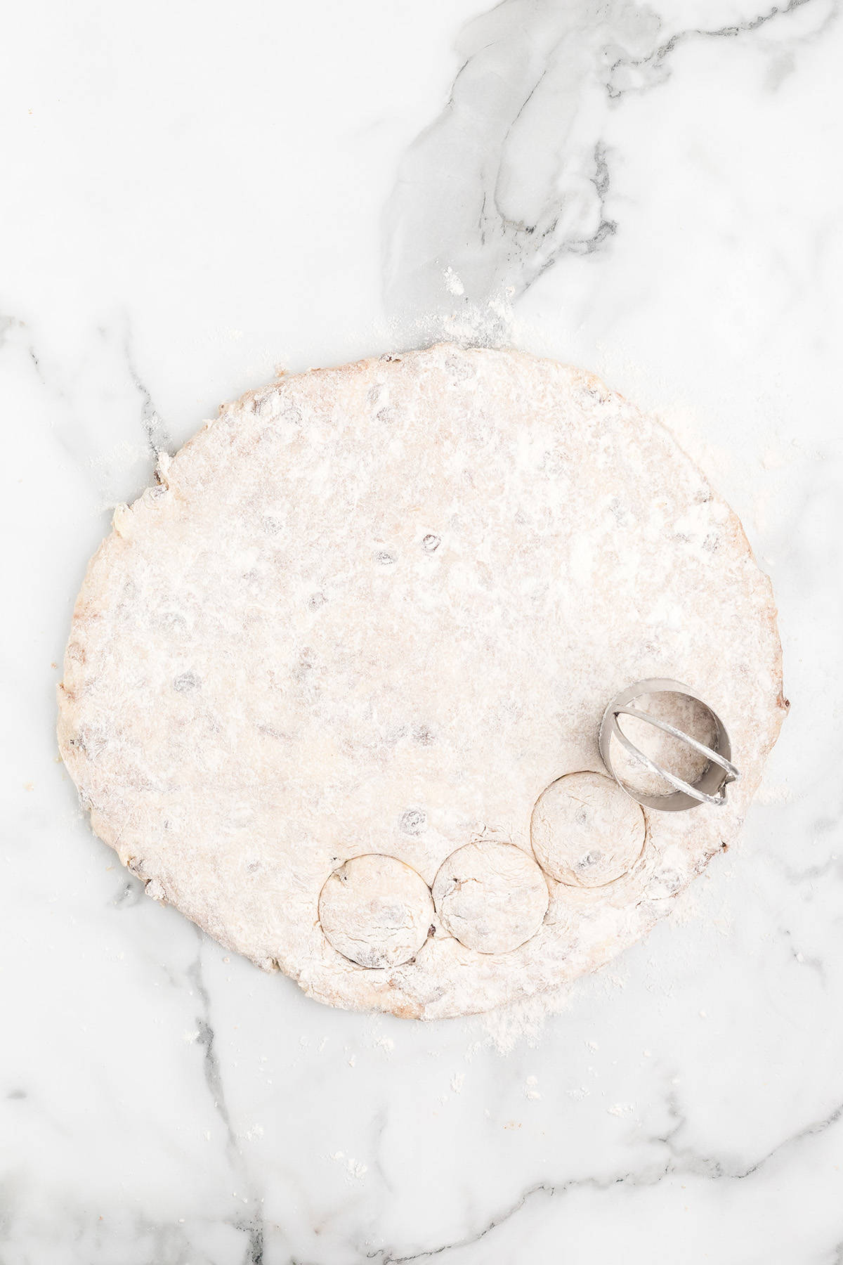 Round biscuit dough on a marble surface with a biscuit cutter making circles along the edge.