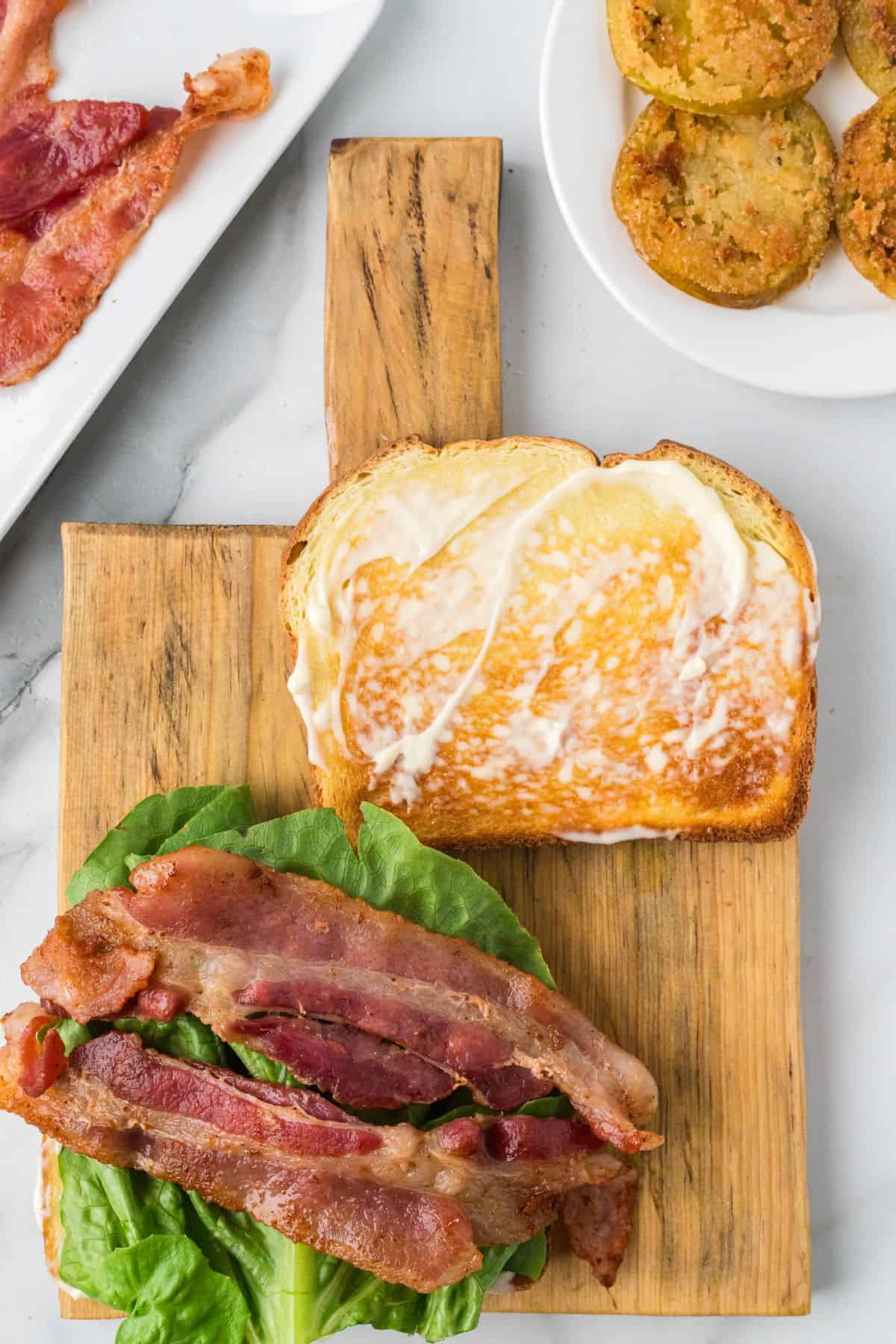 Open BLT sandwich with lettuce and bacon on toasted bread, next to a plate of bacon and fried green tomatoes.