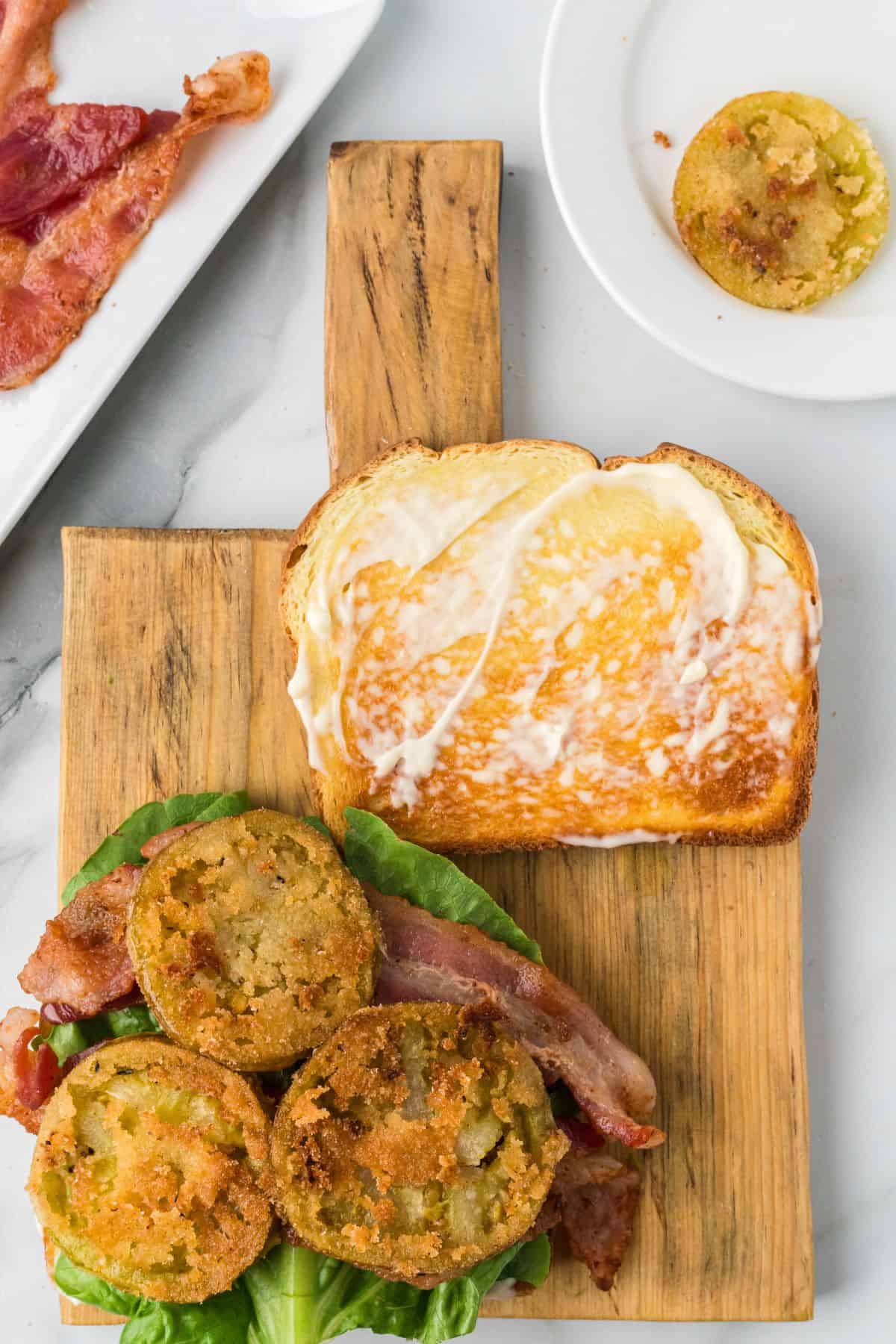 Open sandwich with fried green tomatoes, crispy bacon, lettuce, and mayonnaise on toast, served on a wooden board.