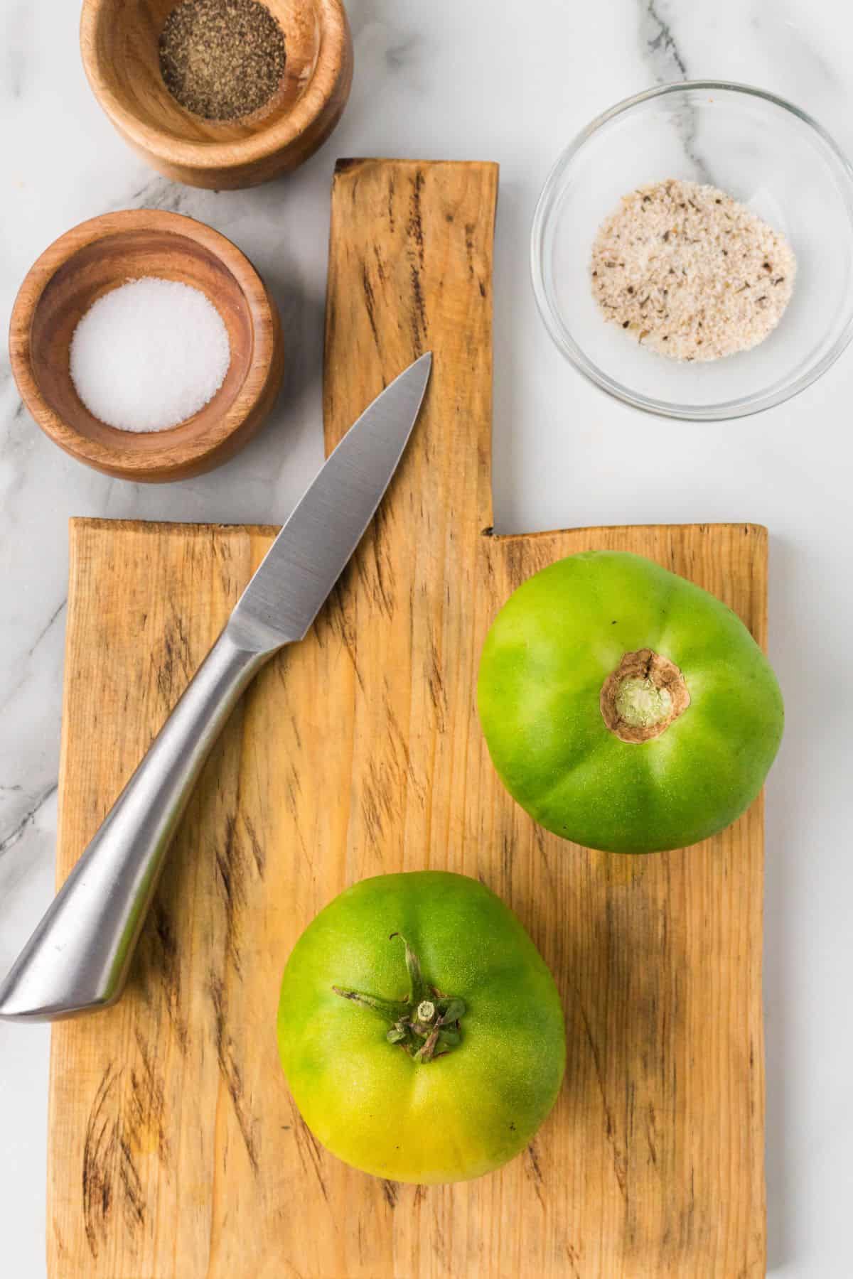 Two green tomatoes and a knife rest on a wooden cutting board, with salt and pepper in small bowls nearby.