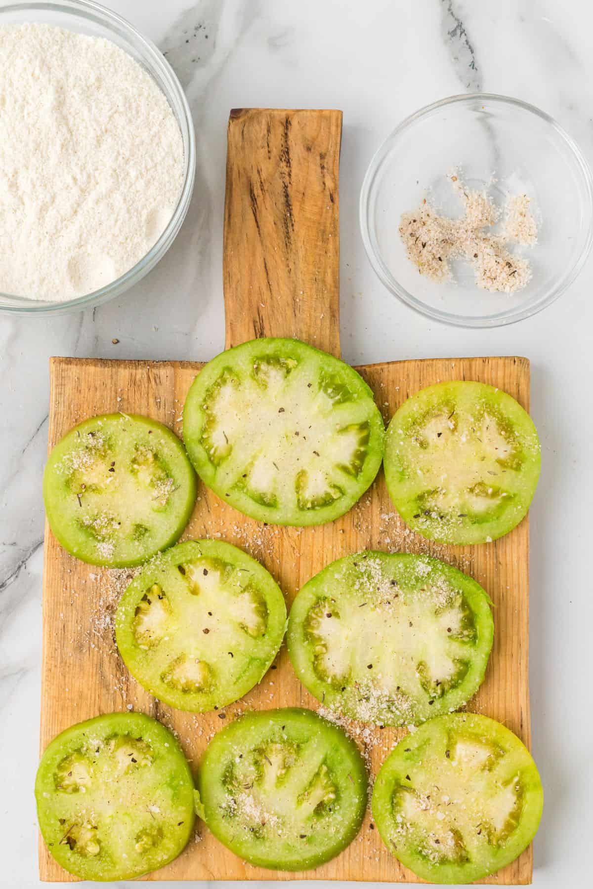 Sliced green tomatoes on a wooden board, with cornmeal and seasoning in bowls on a marble surface.