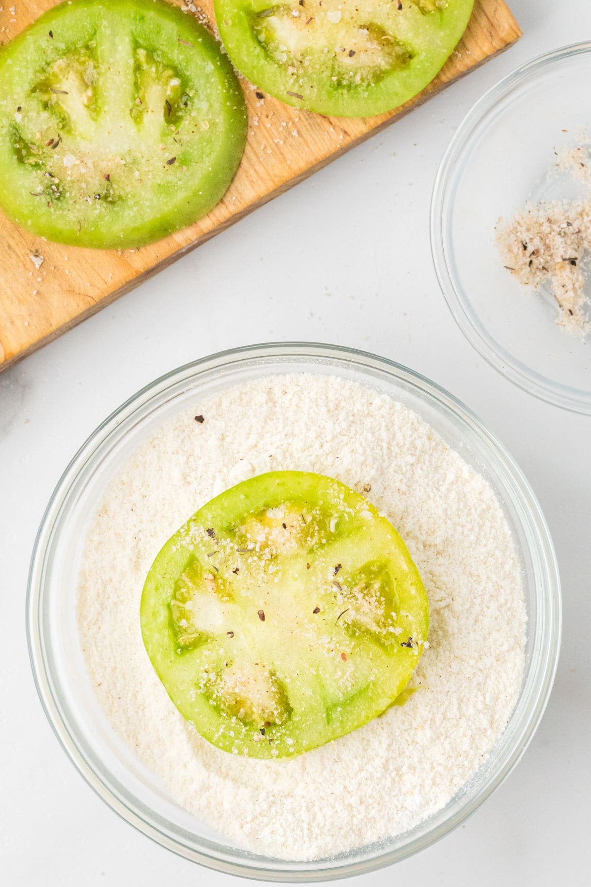 Sliced green tomato in white cornmeal with more slices on a cutting board and a bowl of seasoning nearby.