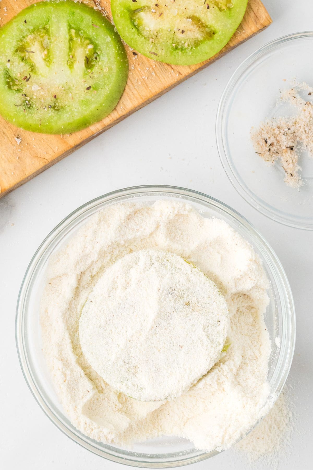 A green tomato slice is being coated in cornmeal, with more tomato slices on a cutting board nearby.