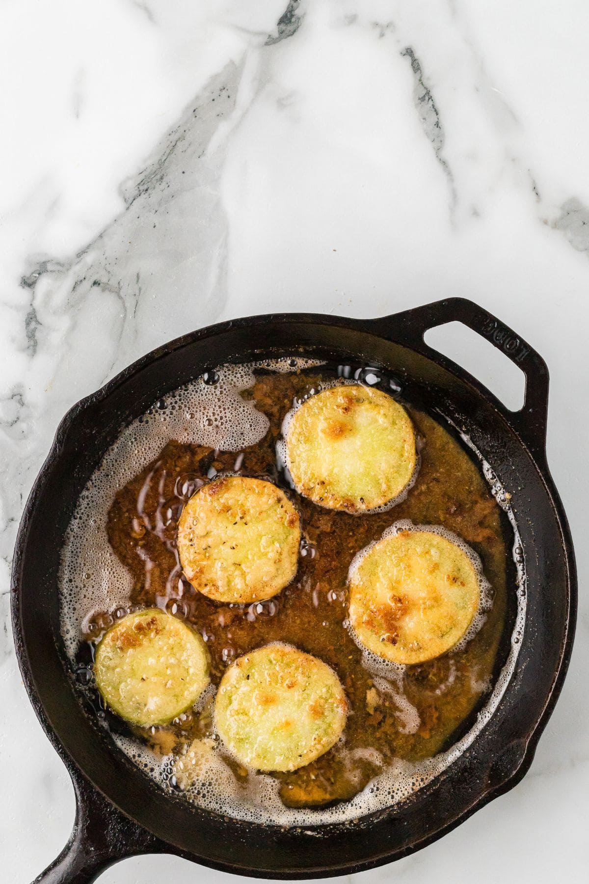 Five slices of breaded tomatoes frying in oil in a black cast iron skillet.