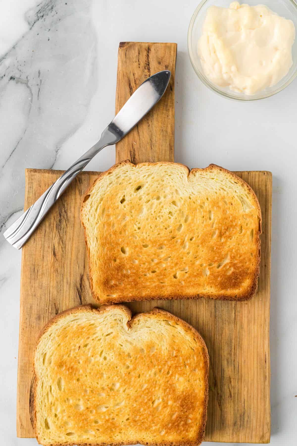 Two slices of toasted bread on a wooden cutting board, with a butter knife and a bowl of mayonnaise nearby.