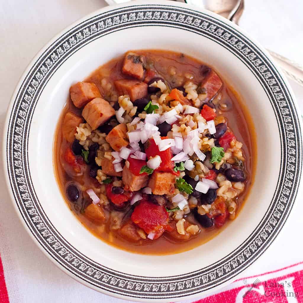 A bowl of soup with diced ham, black beans, tomatoes, rice, and topped with chopped onions and herbs.