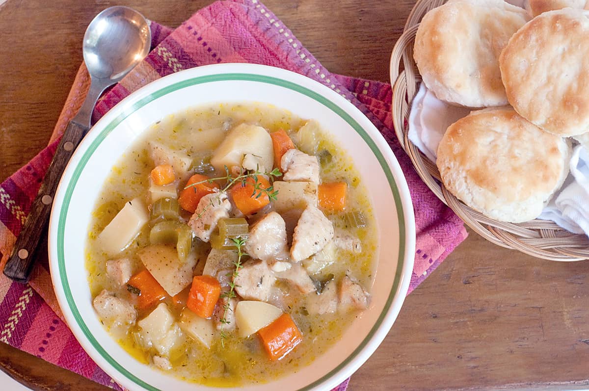A bowl of chicken stew with a basket of biscuits on the side.