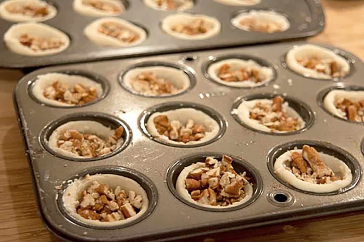 Muffin pans filled with unbaked mini tarts containing chopped nuts on a wooden surface.