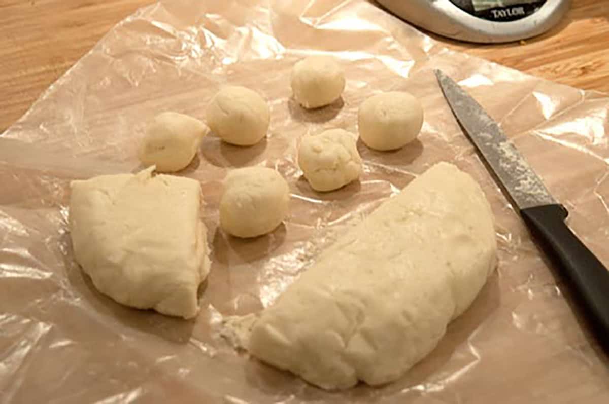 Dough balls and two dough pieces on plastic wrap next to a knife on a wooden surface.