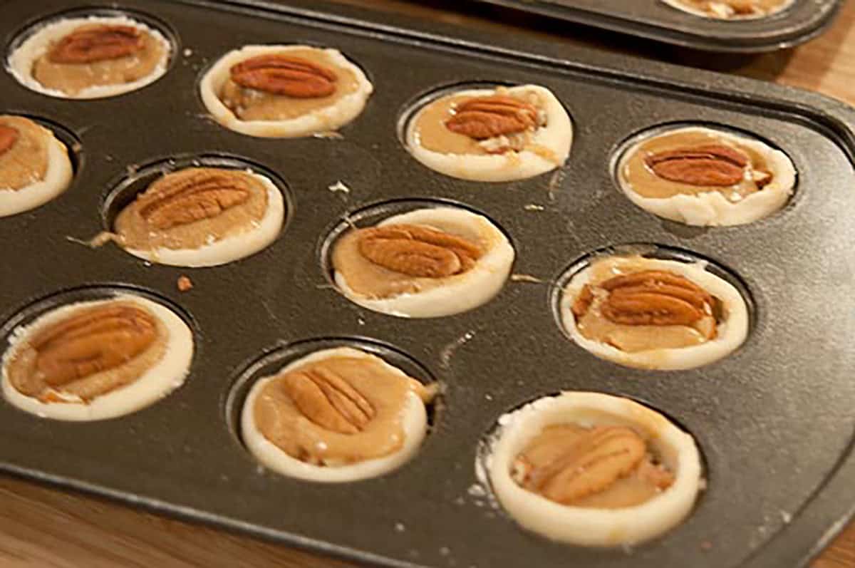 Unbaked pecan tassies in a muffin tin, each filled with pecan topping and topped with a whole pecan.