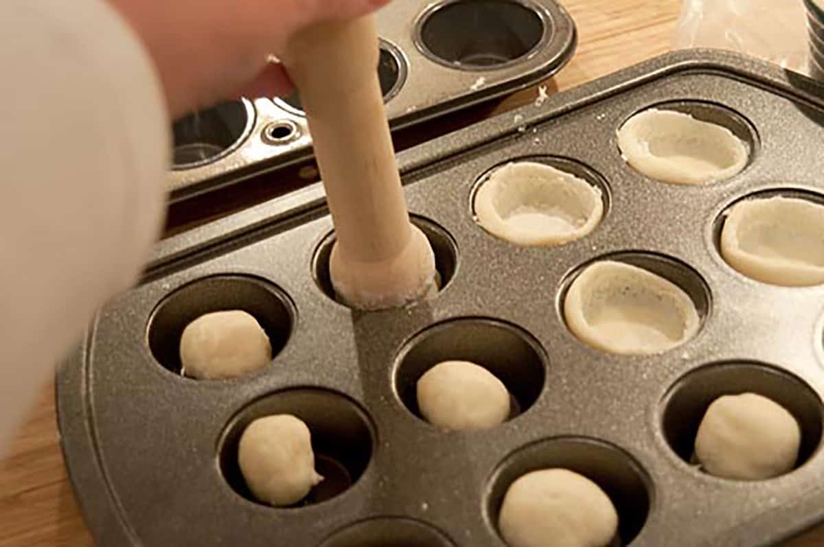 Hand pressing dough into muffin tin cups with a wooden tool, with dough balls in other cups.