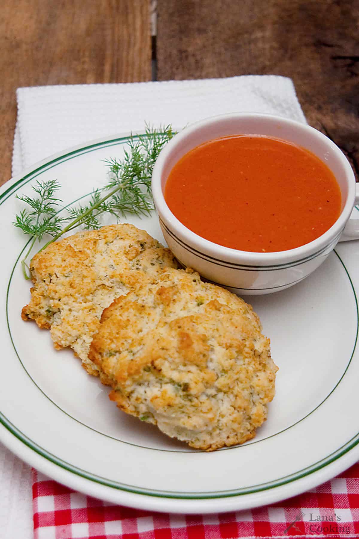 A cup of tomato soup with two biscuits on a plate, garnished with fresh dill, on a wooden table.