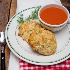 A cup of tomato soup with two biscuits on a plate, garnished with a sprig of fresh dill.