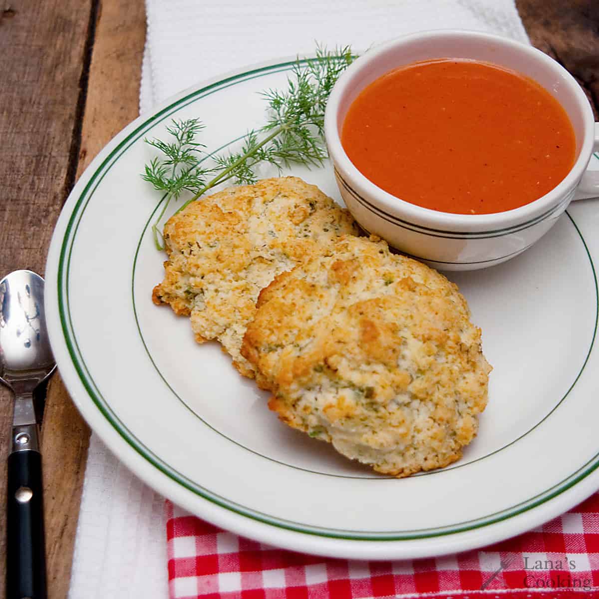 A cup of tomato soup with two biscuits on a plate, garnished with a sprig of fresh dill.
